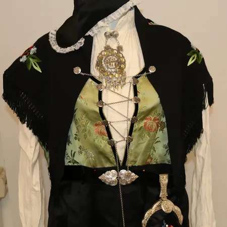 Rogalandsbunad med skjorte, forkle, sjal, veske, belte, hodeplagg, 2 søljer, ett par mansjettknapper, sjalfeste og kjede med spyd. Lengde 136cm, liv 103cm, bryst 112cm. Lite hull ved skjørteåpningen som er sydd sammen med hånd. Vekt: 0 g Kontakt Lånekontoret for frakt