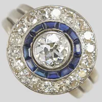 Ring, gammalslipad diamant ca 1,00ct ca Cr(J)/SI1, gammal- och antikslipade diamanter 18xca0,05ct ca W-Ca/VS2-P, syntetiska safirer, millegriffeinfattade, Ø18¼, bredd:4,5-17mm, senare ringskena, spricka i millegriffen, platina/vitguld 18K. Vikt: 10,8 g