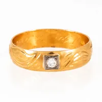 Ring i 23K guld med en diamant c:a 0,08ct; fattad i vitt guld 18K. Mönstergravyr runt om. Den är 5,9 mm bred, är i storlek 20¾ och väger 6,8g. Ringskenan är tillverkad 1904 av EH Yttergren i Umeå. Kattfot. 