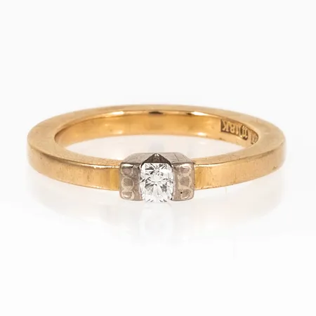 Ring i 18K guld med en kuddslipad diamant c:a 0,20ct (c:a 3,4 x 3,4 mm); fattad i vitt guld. Den är 2,5 - 3,4 mm bred, 22,6 mm hög och är i storlek 17¾. Den väger 6,3g. Tillverkad av Kjellins Guld i Umeå. 