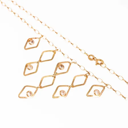 Collier i 18K guld med vita stenar. Den är 42 cm lång, 2,2 mm bred och väger 5,3g. Springring. Den längsta delen i mittpartiet hänger ner 50 mm. 