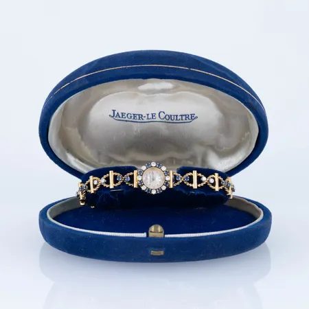 Smyckeur Jaeger LeCoultre i 18K guld med diamanter 14 x c:a 0,02ct & 8 x c:a 0,03ct och 24 safirer. Boett Ø 13,6 mm. Manuellt urverk. Boett no 63670A. Viklås & säkerhetskedja. Innermått stängd c:a 17 cm. Bandet är 8 mm brett. Bruttovikt 20,4g. Originalask. Servicebehov- går sporadiskt- verkar vara dragen i botten. 