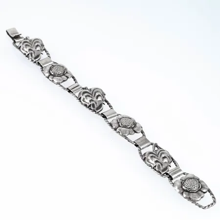 Armband i silver. Dekor av blommor och blad. Det är 19 cm långt, 7,6 - 16,5 mm brett och väger 16,3g. Stämplad E9 (1955), VJ (Victor Janson Guldvaru AB, Lindesberg), Silver & Kattfot. Ojämnheter.