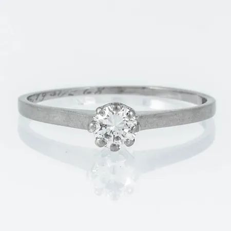 Enstensring i 18K vitguld med en briljantslipad diamant ca 0,30ct. Den är i storlek 18½, skenans bredd 0,8 - 1,8 mm. Vikt: 1,8g. Tillverkad 1963 av Petterssons Eftr Juvelerar AB, Stockholm. Stämplad 18K & Kattfot. Gravyr.