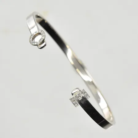 Armring med initialer samt vita stenar, öppen halvstel modell, innermått ca 60x48 mm, oval passform, bredd 4 mm, något skev, vitguld, 18K. Vikt: 20,4 g