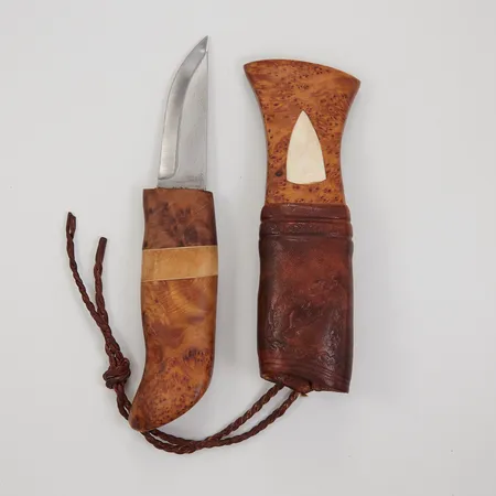 Kniv ca 18cm, skaft i masurbjörk med dataljer i ben, knivblad av Stefan Broström, slidan med monogramsignering A.H (Axel Hansson), viss släppning mellan bendekor och masurbjörk på skaftet 