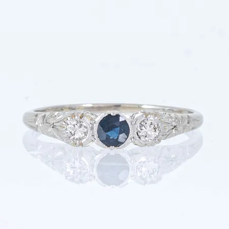 Ring i 18K vitguld med 2 x ca 0,08 ct briljantslipade diamanter, en blå sten (troligen safir). Mönstergravyr. Den är i storlek 19, är 1,1 - 4,2 mm bred och väger 3,1g. Stämplad 18K, svensk importstämpel.