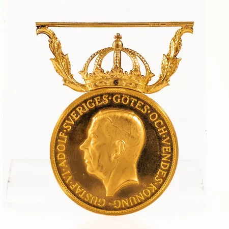 Förtjänstmedalj Gustaf VI Adolf, 23K guld, bandfäste i 18K, Ø 27 mm, vikt: 13,5g. Viktfördelning ca 10,0g i 23K och ca 3,5g i 18K. Personlig gravyr.