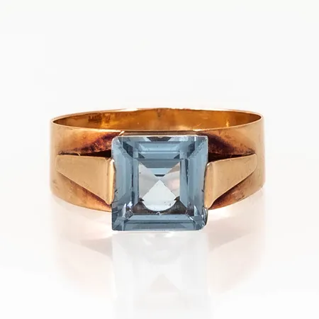 Ring i 18K guld med ljusblå sten 8 x 8 mm. Den är i storlek17¾, är 4,4 - 8 mm bred och väger 2,8g. Stämplad 18K & kattfot. Skena ojämn. Naggade fasetter på sten.