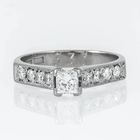 Ring i 18K vitguld med 1 x c:a 0,35ct princesslipad och 8 x c:a 0,03ct briljantslipade diamanter. Den är i storlek 16, är 3 - 5 mm bred och väger 3,7g. Stenstorlek 0,59ct ingraverad. Stämplad AML, 18K. Gravyr. 