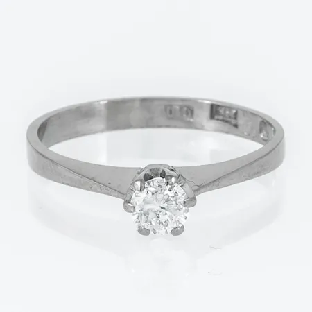 Ring i 18K vitguld med 1 x 0,29ct (c:a W/Pique) briljantslipad diamant. Den är i storlek 16½, är 1,2 x 4,5 mm bred och väger 2,3g. Stämplad G10 (1981), SSG (We-Be Guld AB, Rönninge), 18K & kattfot. Något skev skena.