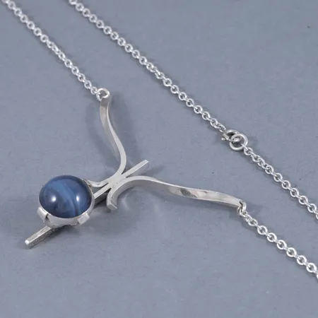 Collier i silver: Integrerat hänge med blå slaggsten. Det är 44 cm långt, 2,5 mm brett och väger 14,5g. Hängets mått 58,5 x 50 mm. Stämplar fattning slaggsten T9 (1969), SOHH (S O Haeggman, Haeggmans Guldsmedsaffär, Nordmaling). Stämplar smyckesdel R9 (1967), VJ (Victor Janson Guldvaru AB, Lindesberg), silver, kattfot. Springring.