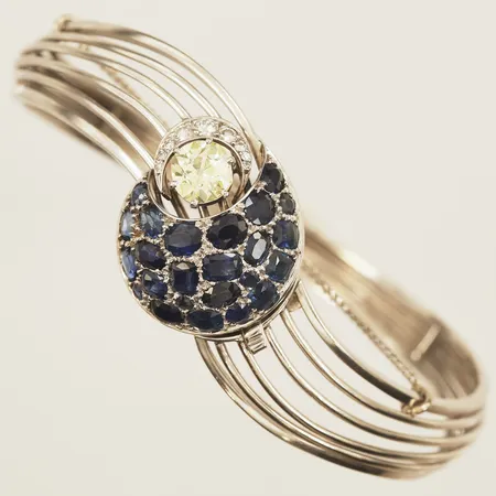 Armring med gammalslipad diamant 1 x ca 1,00ct, ca Yellow(Y-Z)/SI, diamanter 6x ca0,01ct-0,02ct, 3x ca0,03-0,05ct, safirer, innermått ca 18cm, bredd 6,8-21mm, Carlman Aktiebolag Hovjuv, år 1959, säkerhetskedja, vitguld, 18K Vikt: 34 g