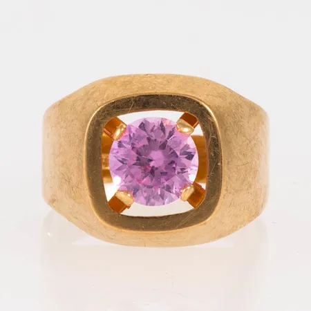 Ring i 18K guld med en rosa sten Ø 7 mm (sannolikt syntetisk rosa safir). Den är i storlek 17¾, är 4 - 14 mm bred och väger 3,6g. Stämplad V9 (1971), HE (Heribert Engelbert AB, Stockholm), 18K och kattfot. 