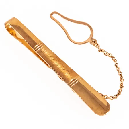 Slipsklämma i 18K guld. Den är 54 mm lång, 4 - 7 mm bred och väger 4,6g. Kedja/spänne i metall. Repor.