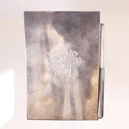 Skrivblock med penna i silver med vapenmonogram som tidigare tillhört Marjorie Merriweather Post (1887-1973) amerikansk affärskvinna, filantrop och societetsdam, arvtagare till C.W Post ägare av General Foods Co, monogrammet med den latinska frasen "In Me Mea Ses Omnis" graverad "Marjorie from ..... March 15th 1957", Marjorie är känd bland annat för bygget av godset Mar-a-Lago, Florida där hon bodde fram till sin död, senare köptes detta av USA:s nuvarande president Donnald Trump, monogrammet på skrivblocket finns fortfarande bevarat i husets eldstad, mått: 8x12cm höjd: 3,1cm, fylld botten med använt pappersblock, engelska stämplar för Gerald Benney London år 1956, silver 925/1000  Vikt: 428,1 g Skickas med postpaket.