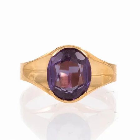 Ring i 18K guld med en lila syntetisk sten 9,5 x 8 mm. Den är i storlek 16½, är 3 - 10,5 mm bred och väger 2,4g. Något skev skena.