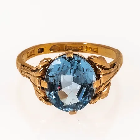 Ring i 23K guld med en blå syntetisk spinell i 18K fattning. Den är 2,2 - 13,4 mm bred, är i storlek 18¼ och väger 4,7g. Ojämn skena med gravyr.