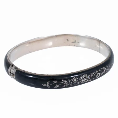 Armring i silver med svart emalj, graverat blommönster och fröpärlor. Innermått 17,5 cm, 50,5 x 58 mm. Den är 9 mm bred och väger 16,0g. Stämplad kattfot.